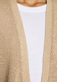 Gros plan d'une personne portant un t-shirt blanc à col rond sous un cardigan beige en tricot avec un motif texturé.