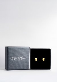 Guldskalleörhängen visade i en låda klädd med svart sammet, inuti en strukturerad svart ytterlåda med vit logotyp på locket.