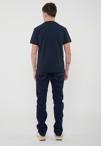 T-shirt navy a maniche corte con dettagli arancioni sulle maniche, abbinato a jeans denim blu scuro e sneakers beige. Design semplice, stile casual.