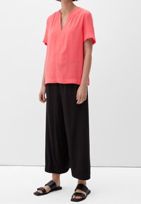 Blouse corail à manches courtes avec un décolleté en V et des détails froncés au niveau de l'encolure, associée à un pantalon noir ample et des sandales plates noires.