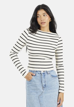 Vrouw met lang donker haar, die gouden creolen draagt, een wit-zwart gestreept shirt met lange mouwen en een lichtblauwe spijkerrok, met haar hand in haar zak.