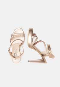 Sandales à talons hauts métalliques dorées avec des brides croisées, une finition brillante, une accentuation par boucle et un talon aiguille pointu.