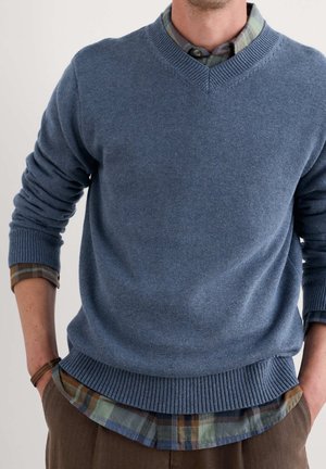 Mann trägt einen blauen V-Ausschnitt-Pullover über einem mehrfarbigen karrierten Hemd mit brauner Hose, Hände in den Taschen, vor einem schlichten Hintergrund.