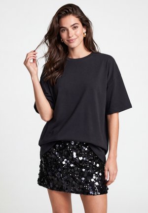 Ung kvinde med langt brunt hår iført en løs sort T-shirt og en sort paillet-mininederdel, smilende og holder en hårtot.