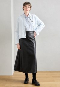 Világoskék steppelt dzseki gombokkal, fehér ing, fekete bőr maxi szoknya és fekete mokaszinek halvány háttér előtt.