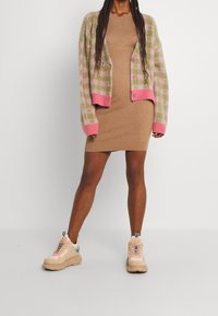 Persona che indossa un vestito a maglia beige a coste, un cardigan a quadri rosa e verde, e sneakers chunky beige, in piedi di fronte a uno sfondo bianco semplice.