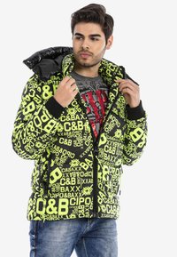 Gepolsterte schwarze Jacke mit neongrüner Markenkennung, ausgestattet mit einem Reißverschluss vorne, hohem Kragen und gerippten Manschetten. Getragen mit einem grauen T-Shirt und blauen Jeans.