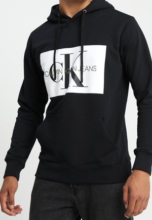 Sweat à capuche noir avec poche avant comportant un grand logo rectangulaire blanc affichant les textes "CK" et "Calvin Klein Jeans".