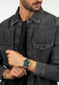 Orologio argento con quadrante verde, indici metallici, cinturino in acciaio inox, indossato con una maglietta nera e una giacca di jeans grigio scuro.