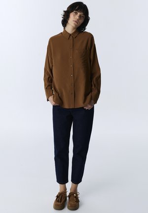 Persona con capelli corti e scuri che indossa una camicia marrone con bottoni, pantaloni scuri corti e scarpe marroni con lacci, in piedi davanti a uno sfondo semplice.