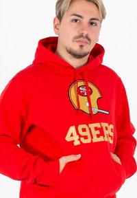 Czerwona bluza z kapturem z dużą grafiką złotej i czarnej hełmu piłkarskiego oraz tekstem "49ERS" w złotym kolorze na piersi. Miękki materiał.