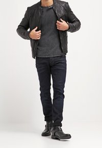 Uomo che indossa una giacca di pelle nera, un maglione grigio a maglia, jeans scuri e stivaletti neri, mentre aggiusta la giacca camminando in avanti.