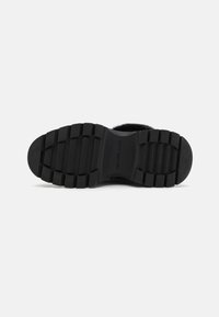 Zwarte rubberen zool met diepe groeven voor grip, met afgeronde randen en een reliëfmerklogo. Zachte, gestructureerde oppervlakte.