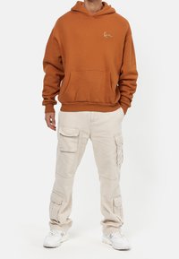 Felpa cappuccio marrone con tasca frontale e logo bianco, abbinata a pantaloni cargo beige con più tasche e vestibilità rilassata.