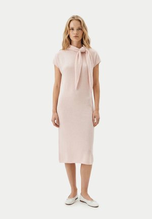 Frau steht und trägt ein hellrosa gestricktes Midikleid mit großer Schleife am Ausschnitt und weißen Ballerinas vor einfachem Hintergrund.