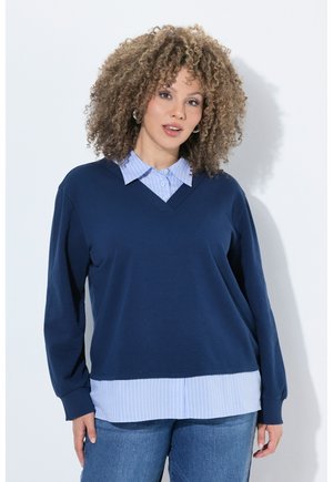 Kvinde med krøllet hår iført en mørkeblå V-hals sweater over en blåstribet skjorte og blå jeans, stående mod en hvid baggrund.