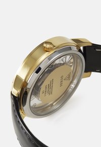 Orologio in acciaio inox color oro con retro trasparente, cinturino in pelle nera e corona. Incisioni notevoli sulla parte posteriore della cassa.