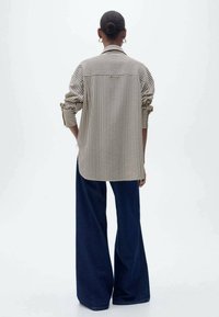 Chemise rayée beige et noire, manches retroussables et coupe décontractée, associée à un pantalon en denim foncé à jambes larges, vue de dos.