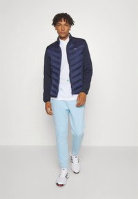 Temno modra puhovka z vzorcem tkanega cik-cak, združena z belo srajco in svetlo modrimi športnimi hlačami. Bele športne superge z sivimi poudarki.