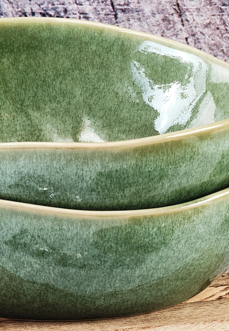 Due ciotole in ceramica impilabili con una sfumatura verde e finitura lucida, bordate di beige, che mostrano una superficie liscia e una forma curvata.