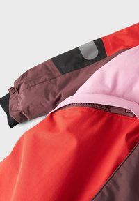 Šareni windbreaker s različitim sekcijama u crvenoj, ružičastoj i smeđoj boji. Ima džep s patentnim zatvaračem i kontrastne crne akcentne rukave.
