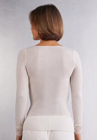 Camiseta ligera de manga larga en suave beige, con una textura lisa y un delicado encaje en los puños. Escote redondeado en la parte posterior.