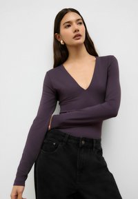 Dlhý rukáv bodysuit v tmavofialovej farbe s hlbokým výstrihom do V. Skombinovaný s čiernymi džínami s vysokým pásom, ktoré majú klasické zapínanie na gombík a vrecká.