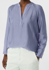 Chemisier lilas à manches longues avec une patte de boutonnage partielle et une coupe décontractée. Le tissu a une finition texturée. Associé à un pantalon blanc.