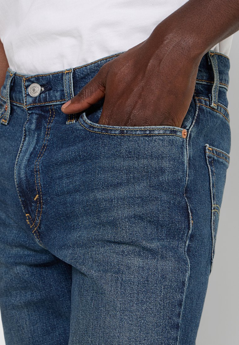 Jean en denim bleu foncé avec une coupe droite, présentant une poche avant et des détails en métal cuivré. Le tissu a une texture lisse.