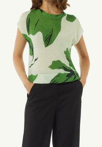 Vihreä ja valkoinen lyhythihainen neulepaita, jossa on lehväkuviointi, teksturoitu neule, pyöreä kaula-aukko ja rento istuvuus, yhdistettynä mustiin housuihin.