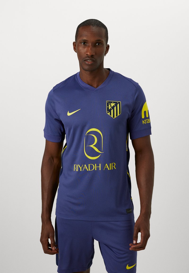 Paarse sporttrui met korte mouwen, gele accenten en een v-hals. Bevat een teamlogo, de tekst "Riyadh Air" en een textuurdesign.