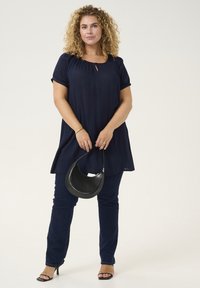 Maglietta blu navy con dettaglio a chiave, maniche corte, abbinata a jeans scuri. Il modello tiene in mano una borsa a mezzaluna nera, indossando tacchi neri.