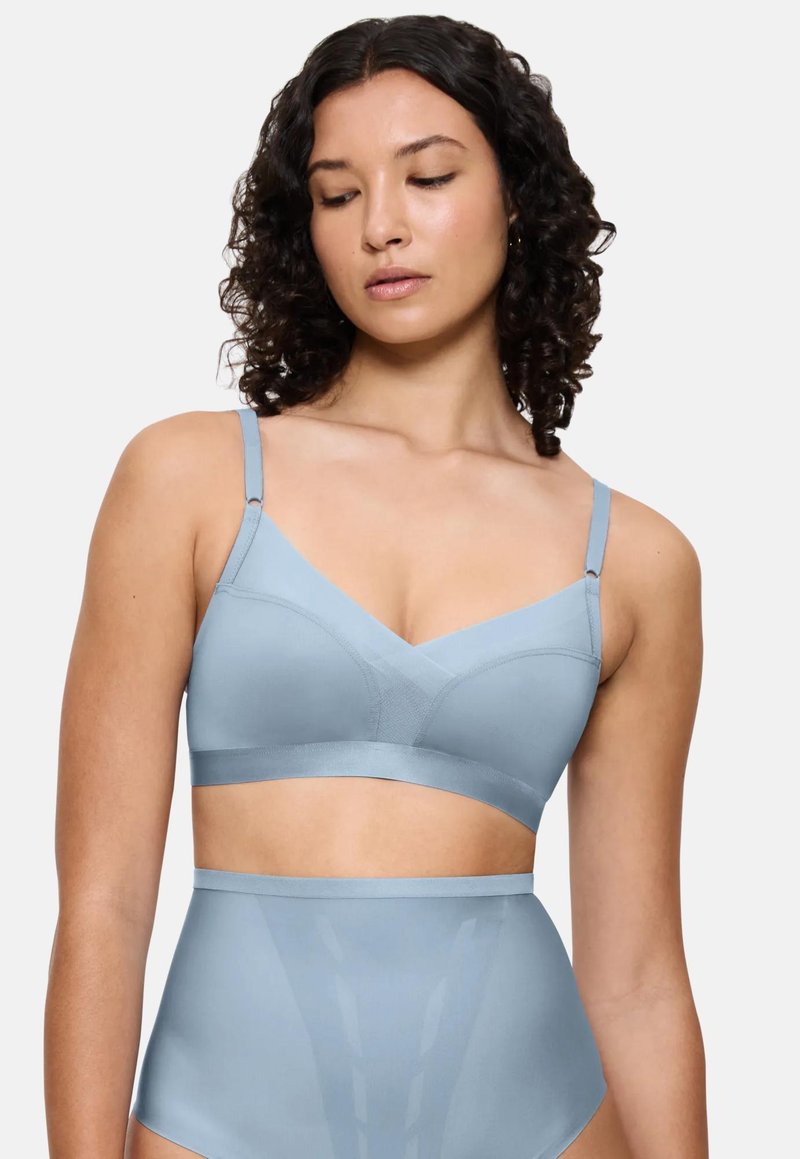 Woman with curly hair wearing a light blue wireless bra and high-waisted shapewear against a plain white background.