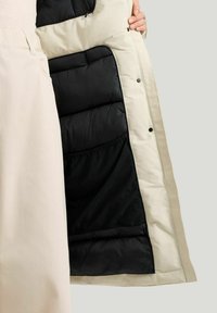 Vinterjacka med en krämfärgad utsida och ett svart kviltat foder. Tyget är slätt med tryckknappar och en strukturerad design.