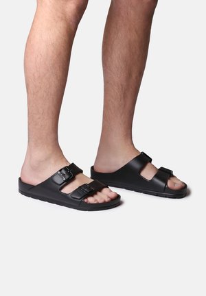 Zwarte schuimslippers van synthetisch materiaal, met twee verstelbare banden met gespen en een open teenontwerp. Getextureerde zool voor grip.
