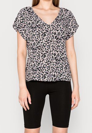 Un haut floral à col V présente un fond noir avec des fleurs blanches et rouges, des manches courtes et une coupe loose, associé à un short de vélo noir.