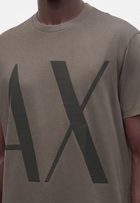 Gros plan d'une personne portant un t-shirt marron avec de grandes lettres foncées "A" et "X" imprimées sur la poitrine.