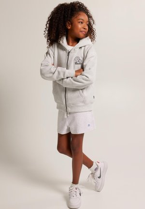 Sweat à capuche gris zippé avec logo, associé à un short gris clair. Le modèle porte des baskets blanches, mettant en avant un style décontracté et confortable.