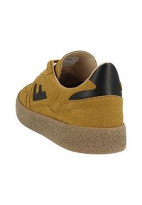 Světlé semišové tenisky žluté barvy s černou koženou patní částí, texturovanou podrážkou a logem. Zaoblený tvar se šněrováním.