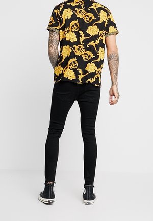 Mężczyzna ubrany w czarne obcisłe jeansy, czarne wysokie trampki i czarną koszulę z krótkim rękawem w żółty kwiatowy wzór, widziany od tyłu.