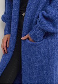 Cárdigan azul de punto con un acabado texturizado, mangas oversized y bolsillos profundos. Lleva encima de un atuendo oscuro, con puños acanalados.