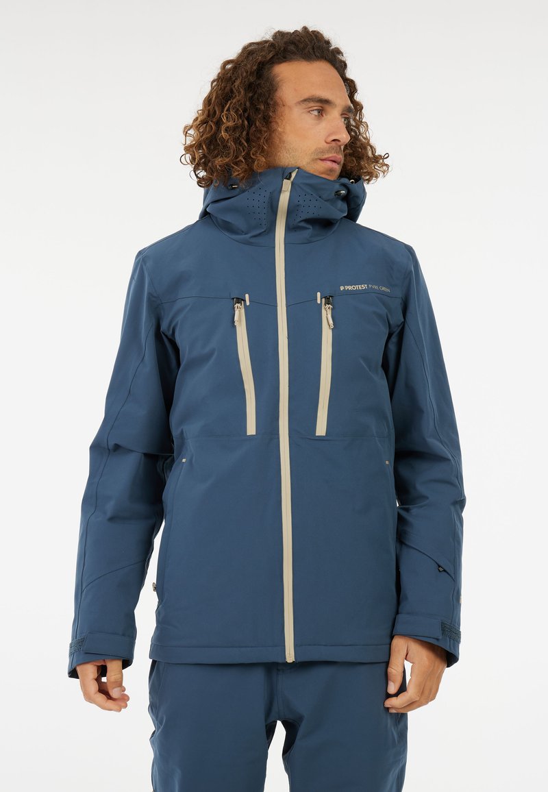 Giacca impermeabile blu con colletto alto, due tasche sul petto con zip e cerniere di un grigio chiaro a contrasto. Dettagli testurizzati sul cappuccio.