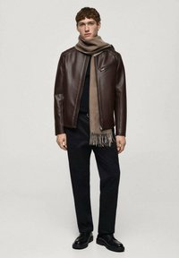 Veste en cuir marron avec une poche zippée, associée à une écharpe beige et un pantalon noir. Texture lisse, design ajusté et style minimaliste.