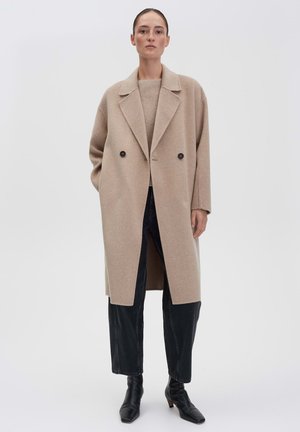 Frau trägt einen beigen, übergroßen Wollmantel über einem passenden Pullover, schwarze, cropped Jeans und schwarze Ankle-Boots mit quadratischer Zehenform. Sie steht vor einem weißen Hintergrund.