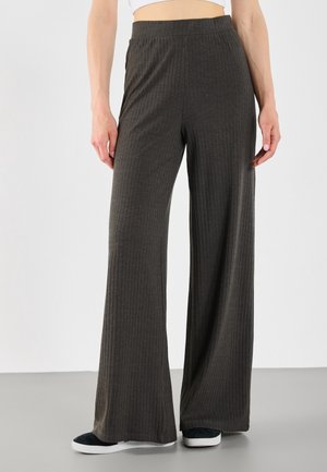 Personne portant un pantalon large côtelé gris foncé et des chaussures noires sans lacets, debout sur un sol gris avec un fond blanc.