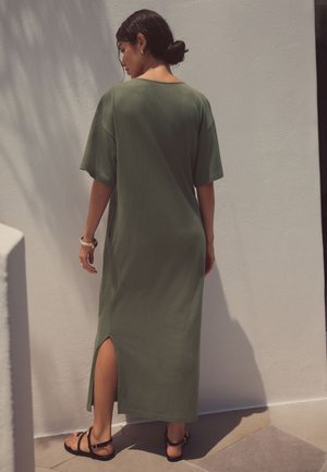 Žena s tmavými vlasy v drdolu, oblečená v zelených midi šatech s rozparkem na zádech a černými sandály, stojící u bílé zdi.