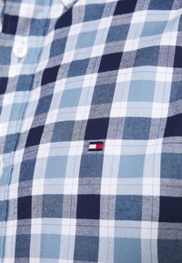Gros plan sur une chemise à carreaux bleue et blanche avec des boutons et un petit logo Tommy Hilfiger brodé sur le tissu.