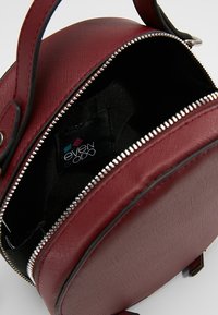 Rund, rød håndtaske lavet af struktureret syntetisk materiale, med lynlåslukning og en sort foret indvendig, hvor et brændemærke er synligt.