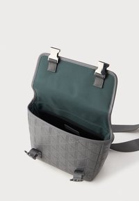 Bolsa transversal cinza com padrão geométrico em relevo, aba aberta com dois fechos metálicos e forro interior em tecido azul-petróleo, apresentada em fundo branco.