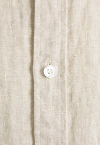 Gros plan sur un tissu en lin beige avec une couture verticale et un bouton blanc à deux trous cousu sur la couture.